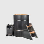 The Star Treatment 2.0 Cold Plunge features a tall, black cylindrical wooden tub with metal bands, a premium cooling unit on wheels, and a set of three metal and wood steps on the right, all set against a plain light gray background.
