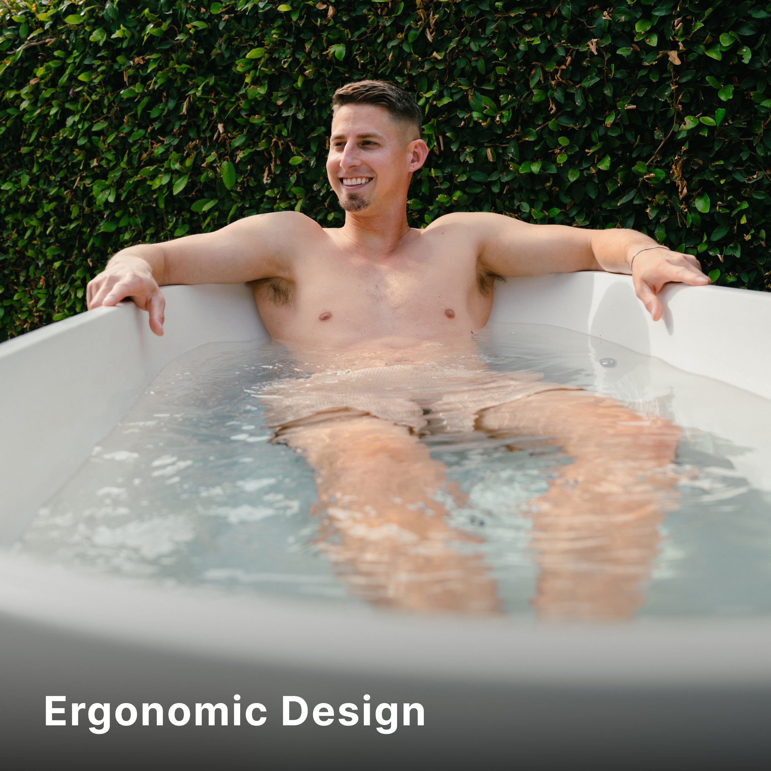 A smiling man relaxes in an outdoor bathtub filled with water. He is sitting comfortably with his arms resting on the tub, and lush green foliage is in the background. Text reads Ergonomic Design at the bottom.
