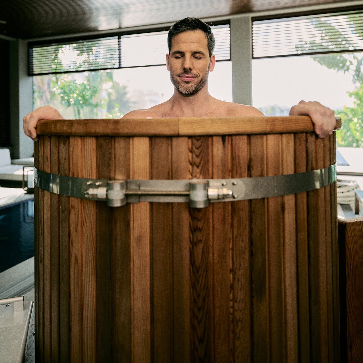 A man with short dark hair stands in a wooden tub filled with water, eyes closed and looking relaxed. Natural light filters through large windows in the background.