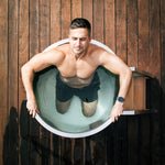 A man with short dark hair and a smartwatch kneels in a Cyber Barrel Commercial Cold Plunge on a wooden deck, eyes closed and hands gripping the sides of the stainless steel tub for optimal recovery.