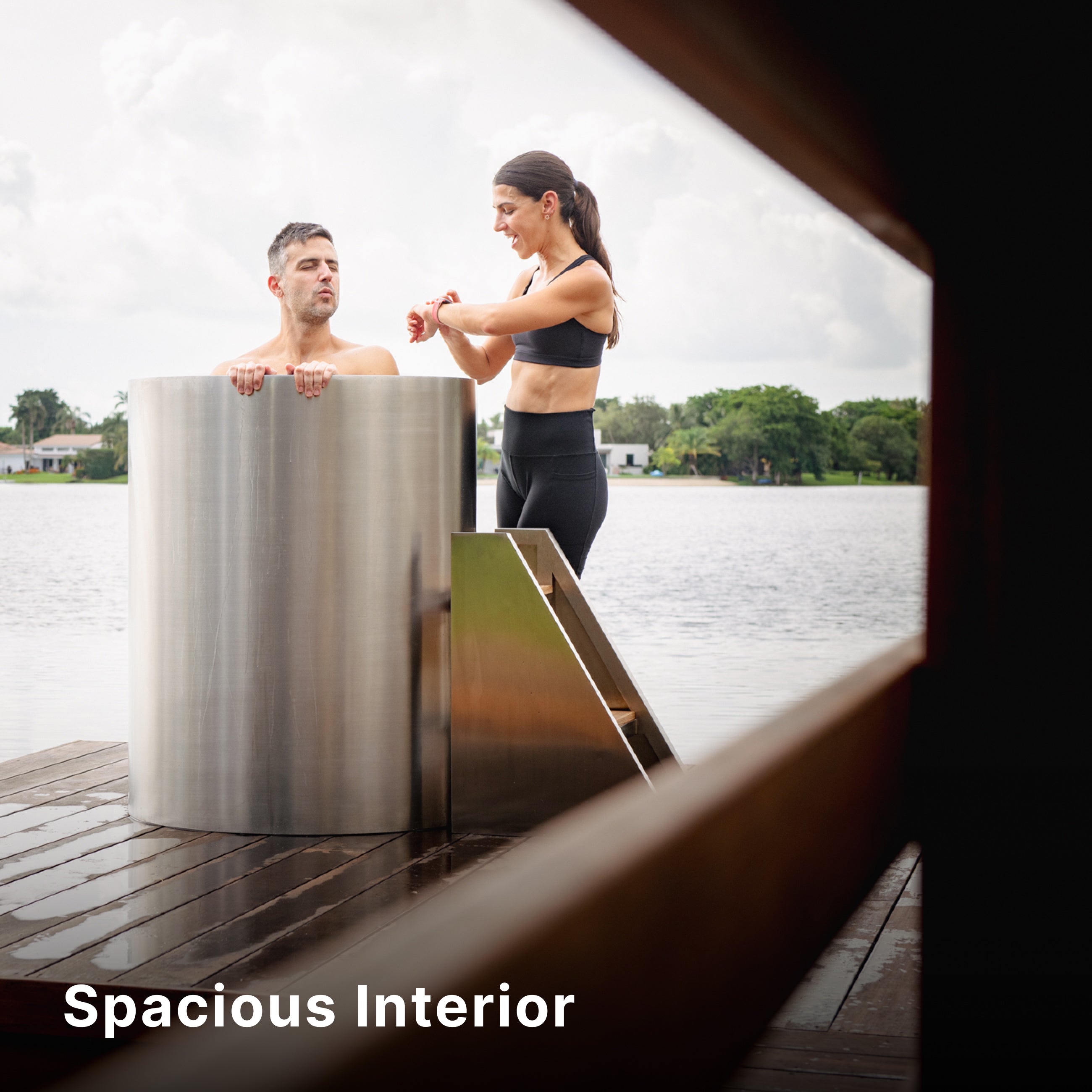 A man stands in a sleek, stainless steel ice bath on a dock by a lake, while a woman in athletic wear checks her watch next to him. The words Spacious Interior appear in the lower left corner.