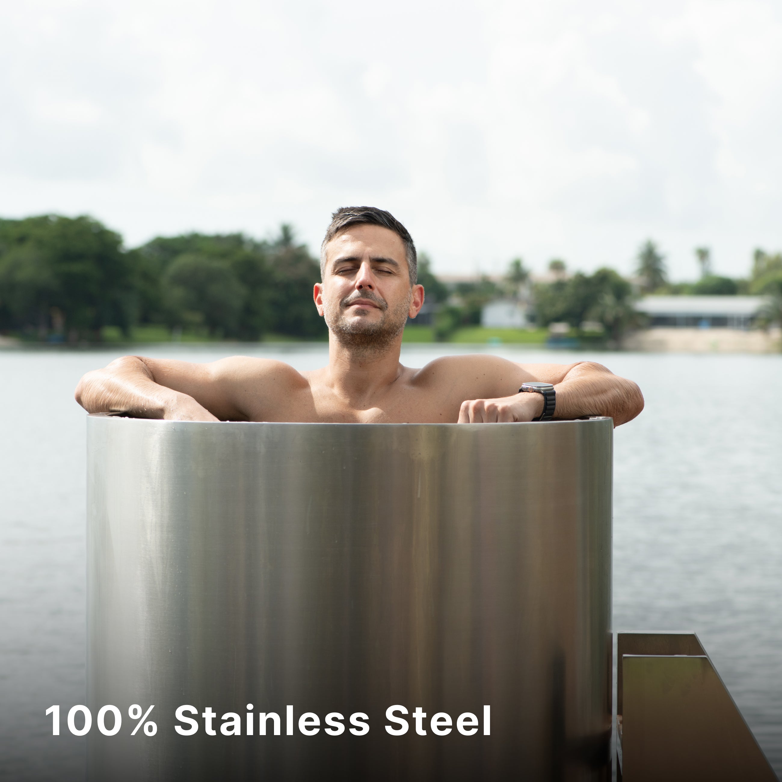 A man relaxes with his eyes closed while sitting in a tall, cylindrical stainless steel tub by a lake. Trees and buildings are visible across the water. Text reads, 100% Stainless Steel.
