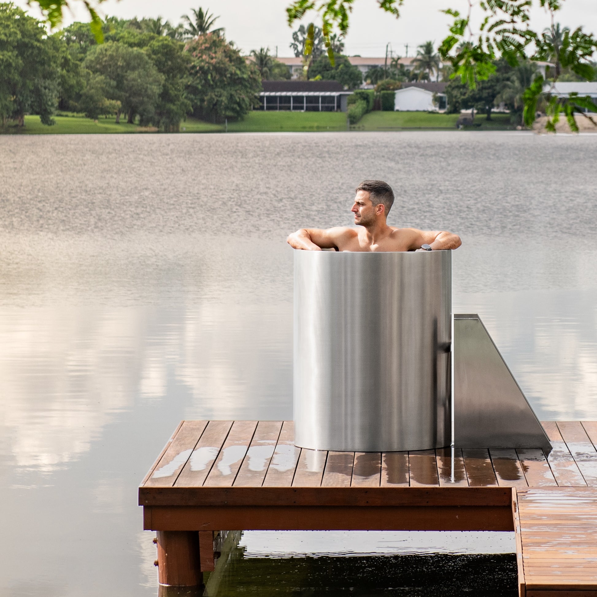 A man relaxes in a tall, cylindrical metal tub on a wooden dock overlooking a calm lake, with trees and houses visible in the background.