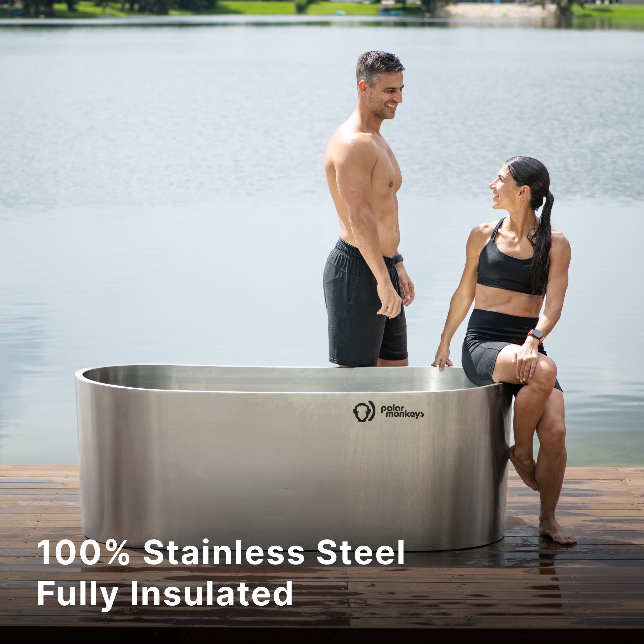 A man and a woman in athletic wear stand beside and sit on the edge of a large stainless steel tub on a dock by a lake. Text reads: 100% Stainless Steel Fully Insulated.