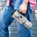 Dressed in blue jeans and a pink shirt, someone stands by a gray textured wall holding a Polar Monkeys Tumbler with straw and handle, showcasing a black lion design—perfect for staying hydrated.