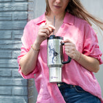 A woman in a loose pink shirt holds a Polar Monkeys Tumbler with handle and straw, showing a wizard riding a cat. She stands by a gray brick wall, ready for serious hydration with her stylish stainless steel Polar Monkeys Tumbler.