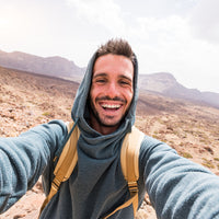 A smiling person wearing a hooded sweatshirt and backpack takes a selfie in a rocky, mountainous landscape under a bright sky.