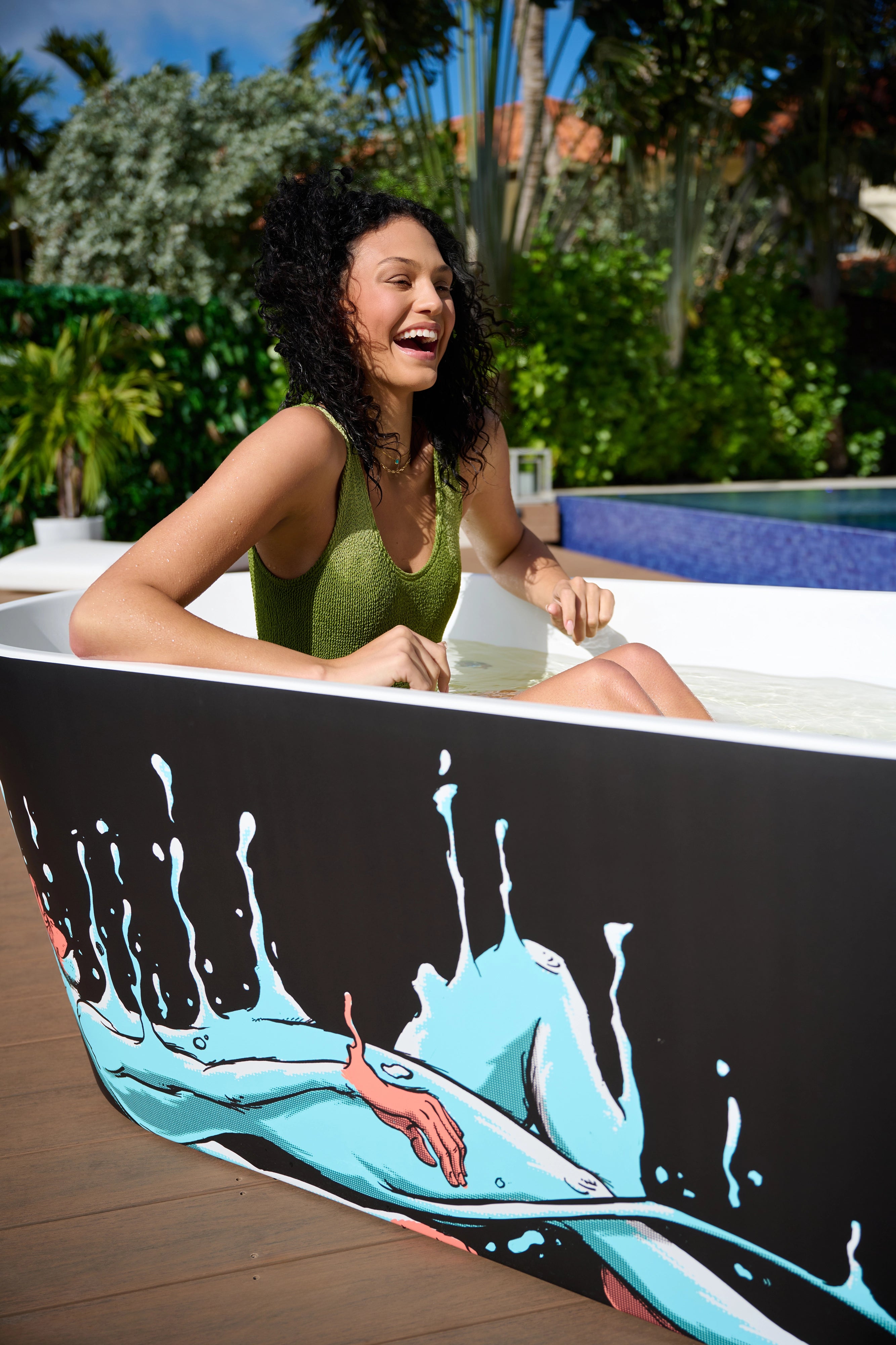 A woman with curly hair wearing a green swimsuit sits smiling in a white bathtub with a black exterior decorated with a colorful illustration, outdoors near a pool and lush greenery.