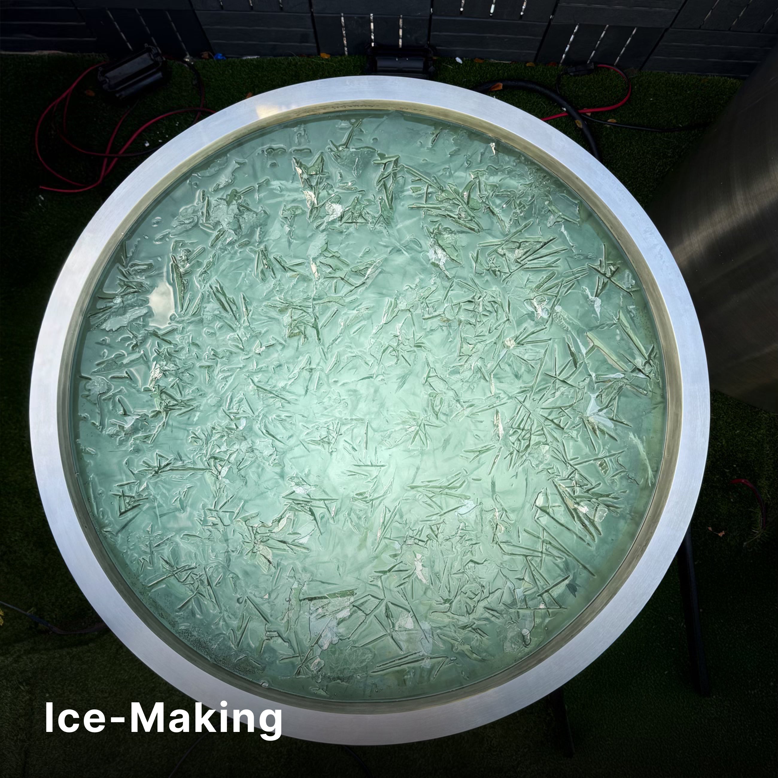 A round metal container filled with water and thin, jagged ice forming on the surface. The background features green artificial grass and a dark fence. The text Ice-Making appears in the lower left corner.