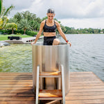 A woman in athletic wear steps into the Cyber Barrel Cold Plunge, a marine-grade stainless steel tub, set on a wooden dock by a lake with lush greenery and palm trees beneath a partly cloudy sky.