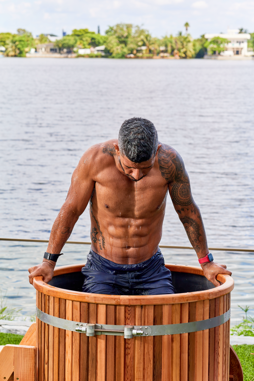 A muscular man with tattoos is emerging from a wooden ice bath outdoors, wearing dark shorts and smartwatches. A lake and trees are visible in the background under a partly cloudy sky.