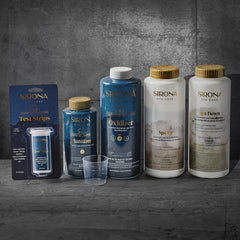 The 6 Month Sanitation Kit, featuring six Sirona Spa Care products—including sanitizer and test strips—is arranged on a gray stone surface with a matching background. A small plastic cup sits in front, ideal for spa or cold plunge use.