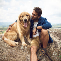 A man sits on a large rock outdoors, smiling and petting a happy golden retriever with its tongue out. The background shows a natural landscape with trees and sky.