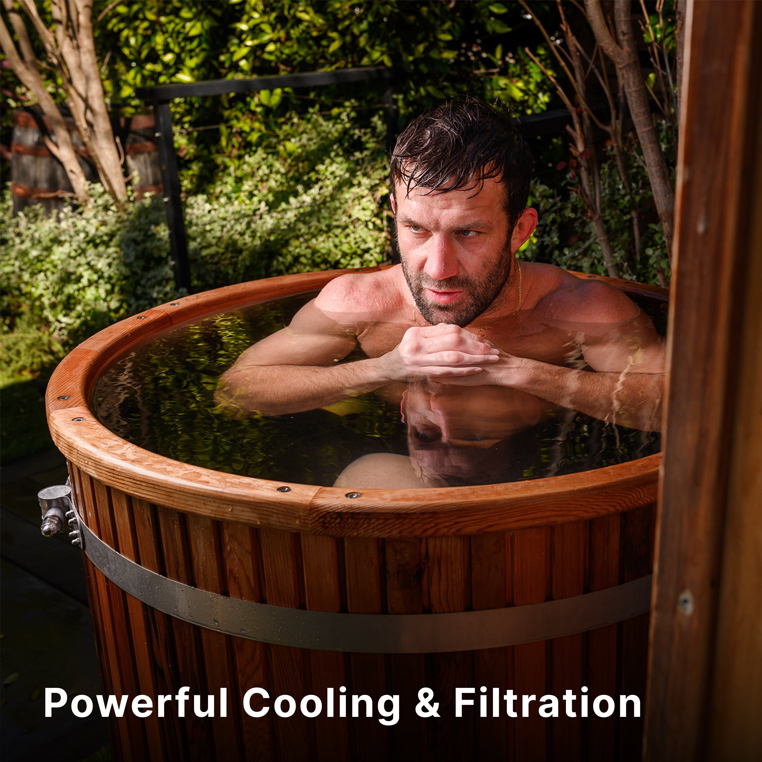 A man is sitting in an outdoor wooden cold plunge tub filled with water, surrounded by greenery. Text at the bottom reads, Powerful Cooling & Filtration.