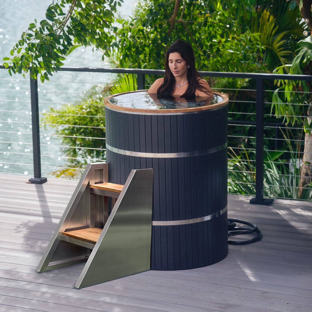 A woman relaxes in the Star Treatment 2.0 Cold Plunge, a premium tall black outdoor soaking tub with metal bands on a wooden deck by the water—ideal for cold plunges or unwinding in compact spaces, surrounded by serene trees and greenery.