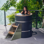 A woman relaxes in the Star Treatment 2.0 Cold Plunge, a premium tall black outdoor soaking tub with metal bands on a wooden deck by the water—ideal for cold plunges or unwinding in compact spaces, surrounded by serene trees and greenery.