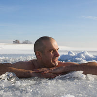 A man with a shaved head is relaxing in icy water outdoors, leaning on the edge of the ice with his arms folded, surrounded by snow and ice under a clear blue sky.
