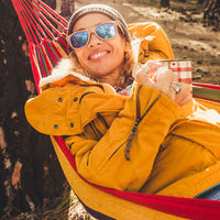 A smiling person in a yellow jacket and sunglasses relaxes in a colorful hammock, holding a mug, with sunlight filtering through trees in the background.