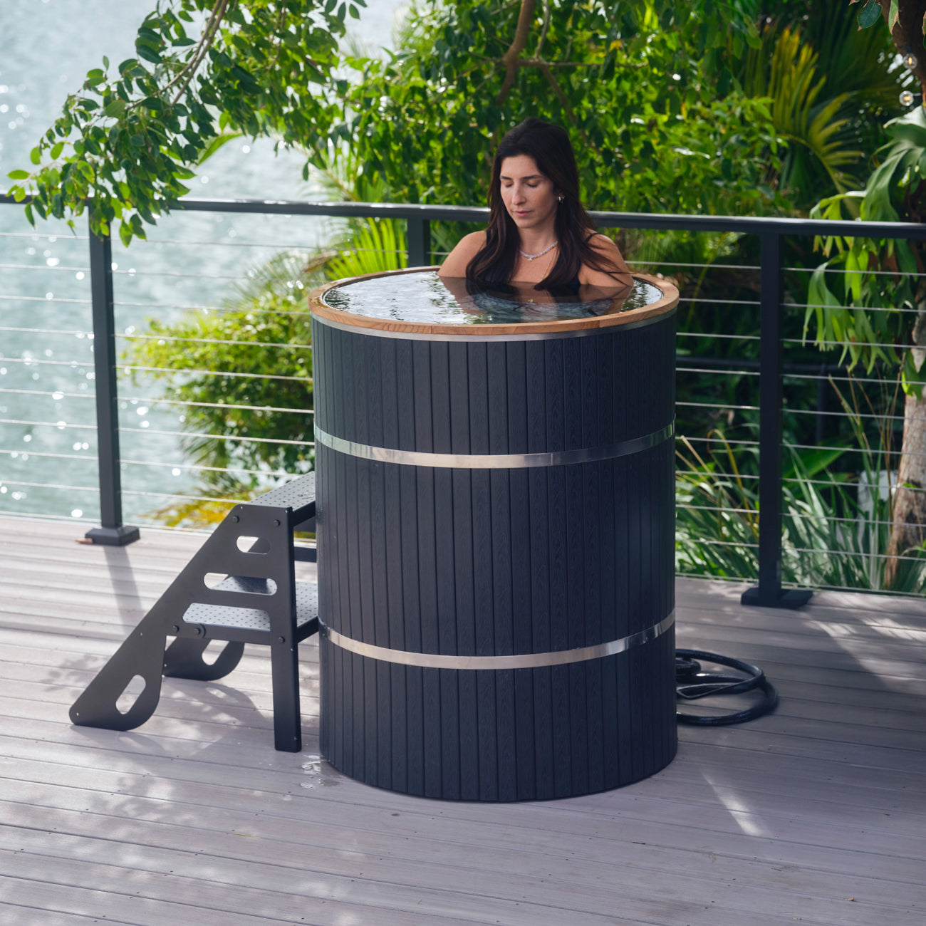 A woman enjoys the Star Treatment 2.0, a premium tall black cylindrical outdoor soaking tub on a wooden deck with water views—ideal for cold plunge therapy or relaxing in compact spaces, featuring a black metal step ladder.