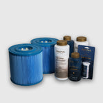 Two blue cylindrical cold plunge filters sit next to bottles and packets from the 6 Month Care Kit, which includes a chlorine-free sanitizer, test strips, and other water treatment chemicals, against a plain light background.