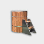 The Star Treatment 2.0 Commercial Cold Plunge, a cylindrical wooden tub with metal bands for cold therapy, is shown beside compact stairs with wood steps and metal sides, set against a plain light background.