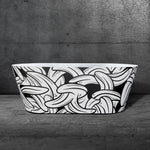 The Brainpod 2.0 Commercial, a modern freestanding bathtub featuring an ergonomic design and a black exterior with a bold white banana pattern, sits on a gray stone floor against a textured dark gray wall.