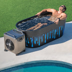 A man wearing sunglasses relaxes in a Brainpod 2.0 Cold Plunge tub with blue drips, filled with water, beside a pool. A cooling fan is set up next to the tub on the pool deck.