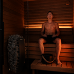 A man sits calmly on a wooden bench inside the Baia Glass Sauna, wearing black shorts. Sauna stones and a water bucket are visible nearby, with warm orange lighting highlighting the wooden interior.