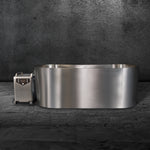 The Cyber Plunge, a modern metallic oval bathtub, sits on a dark textured floor against a matching wall. Next to it is a wheeled machine with controls and handle, all crafted from marine-grade stainless steel for durability and style.