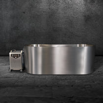 The Cyber Plunge Cold Plunge, a modern freestanding marine-grade stainless steel bathtub, sits on a concrete floor by a dark textured wall, paired with a compact metallic wheeled heater unit for a distinctive and stylish look.