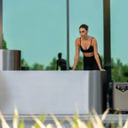 A woman in black athletic wear leans on a modern, metallic counter outdoors, framed by glass windows reflecting greenery—an ideal recovery environment inspired by Contrast Edition dual orientation therapy.
