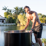 A shirtless man in black shorts stands beside a Cyber Barrel Commercial cold plunge tub outdoors, one hand on its edge. Palm trees, water, and a house in the sunny background set the scene for optimal recovery in style.