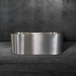 The Cyber Plunge Commercial, a modern freestanding bathtub made of marine-grade stainless steel, sits on a gray marble floor with a textured dark gray wall behind it.