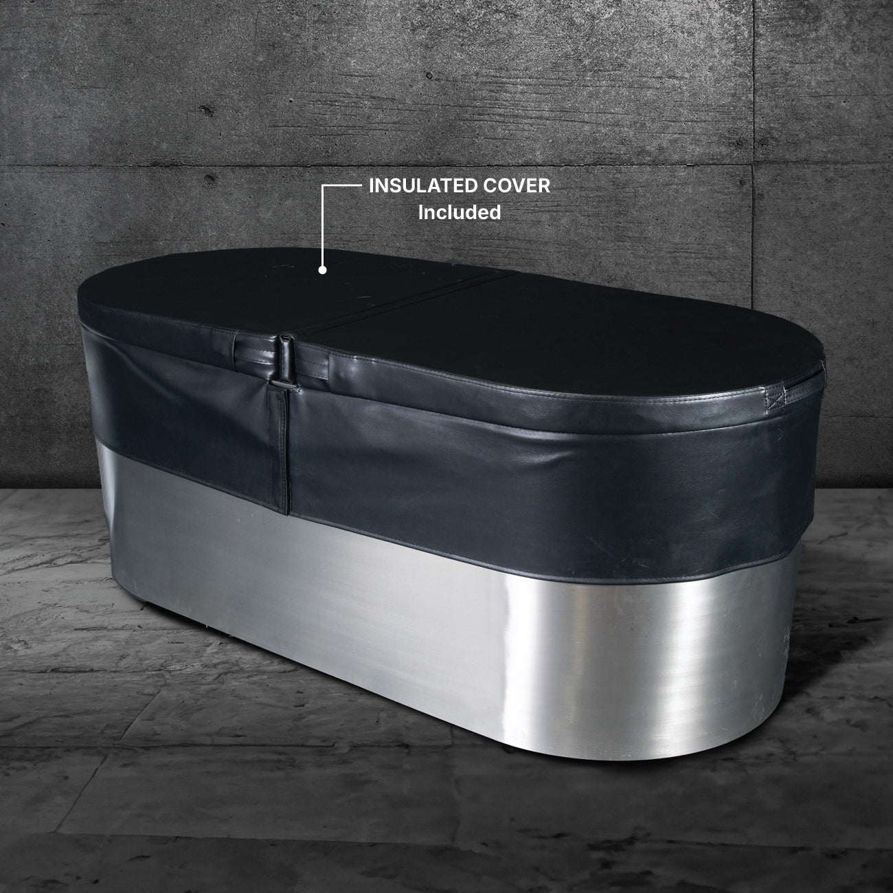 The Cyber Plunge, a marine-grade stainless steel oval tub with a black insulated cover (included), sits on a gray stone floor against a textured concrete wall, creating a distinctive look.