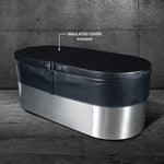 The Cyber Plunge, a marine-grade stainless steel oval tub with a black insulated cover (included), sits on a gray stone floor against a textured concrete wall, creating a distinctive look.