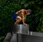 A shirtless man in blue shorts climbs into the large stainless steel Contrast Edition ice bath outdoors, surrounded by lush green plants and trees—an ideal recovery environment for those utilizing contrast therapy systems.