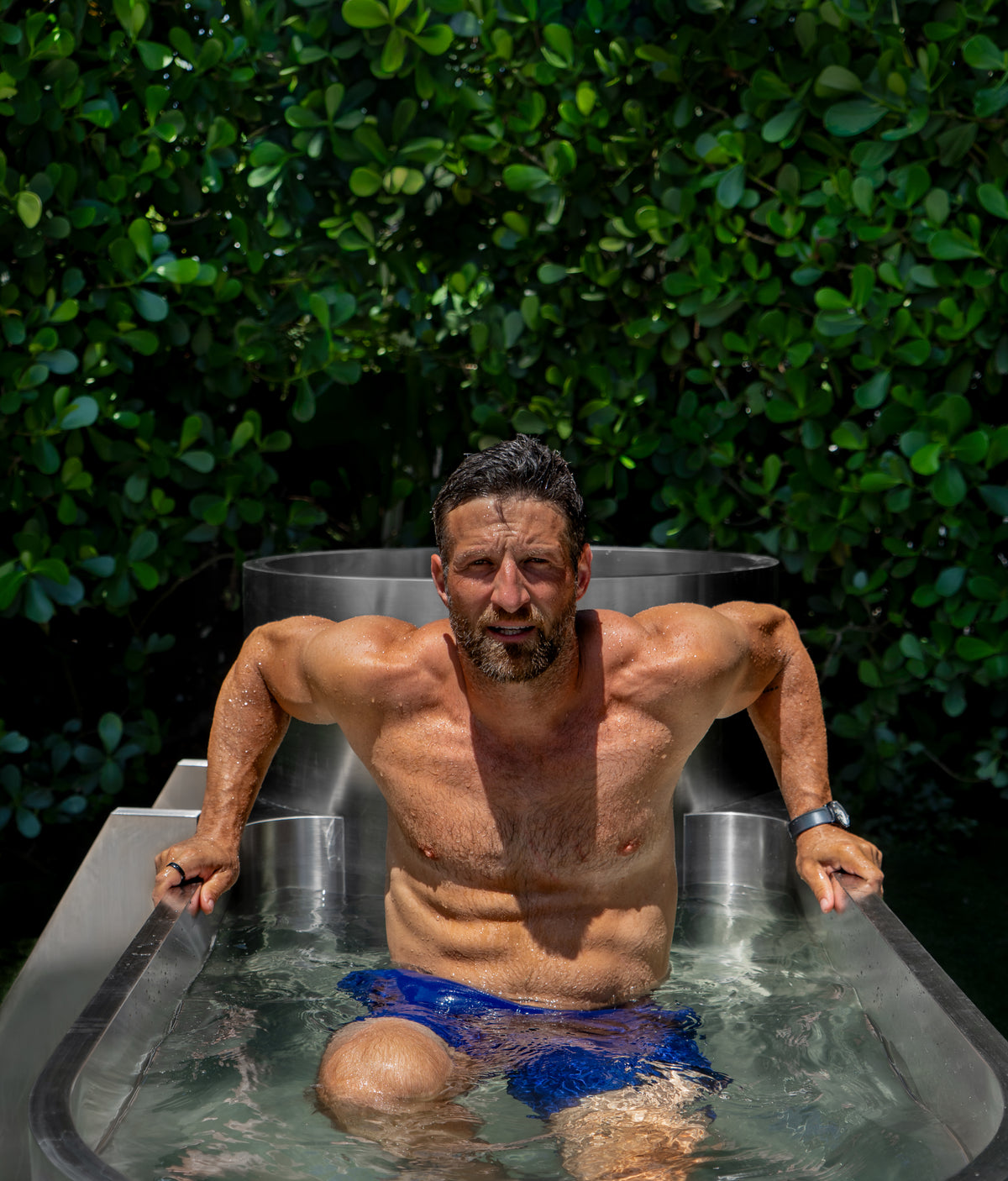 A muscular man in blue swim trunks is emerging from the Contrast Edition, an outdoor metal tub filled with water and surrounded by lush green foliage, creating the perfect recovery environment for hot and cold therapy.