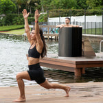 A woman in athletic wear lunges on a dock by the water as a man enjoys optimal recovery in a Cyber Barrel Commercial marine-grade stainless steel tub; trees and a fence are visible in the background.
