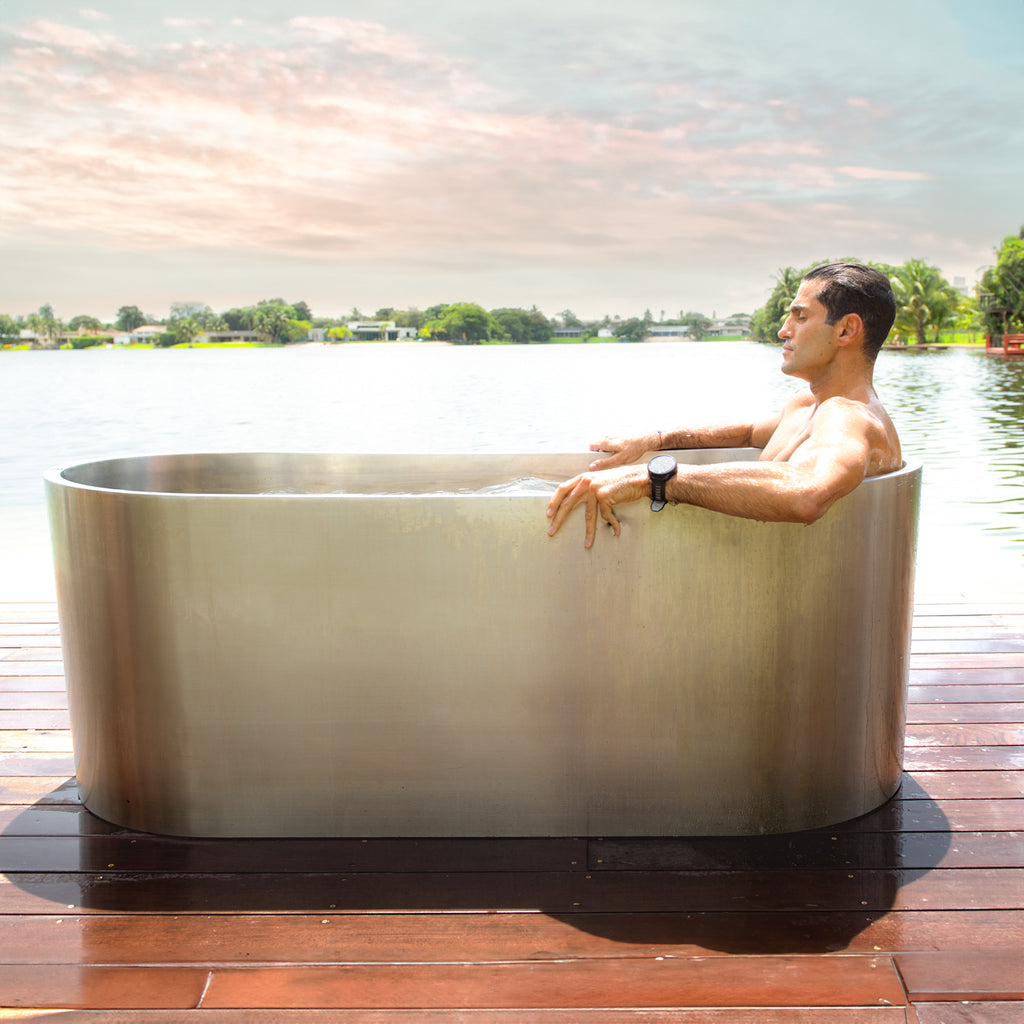 A man relaxes in a Cyber Plunge Cold Plunge, an oval marine-grade stainless steel tub, on a wooden deck by a lake with trees and a colorful sky in the background.