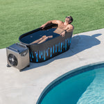 A man wearing sunglasses relaxes in a Brainpod 2.0, an artist-designed acrylic cold plunge with blue and white drips, on a poolside patio next to a portable cooling unit, with grass visible in the background.