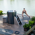 A woman in workout attire does side lunges with dumbbells on a wooden deck beside a Cyber Barrel cold plunge tub by a lake, surrounded by green plants and fitness equipment.