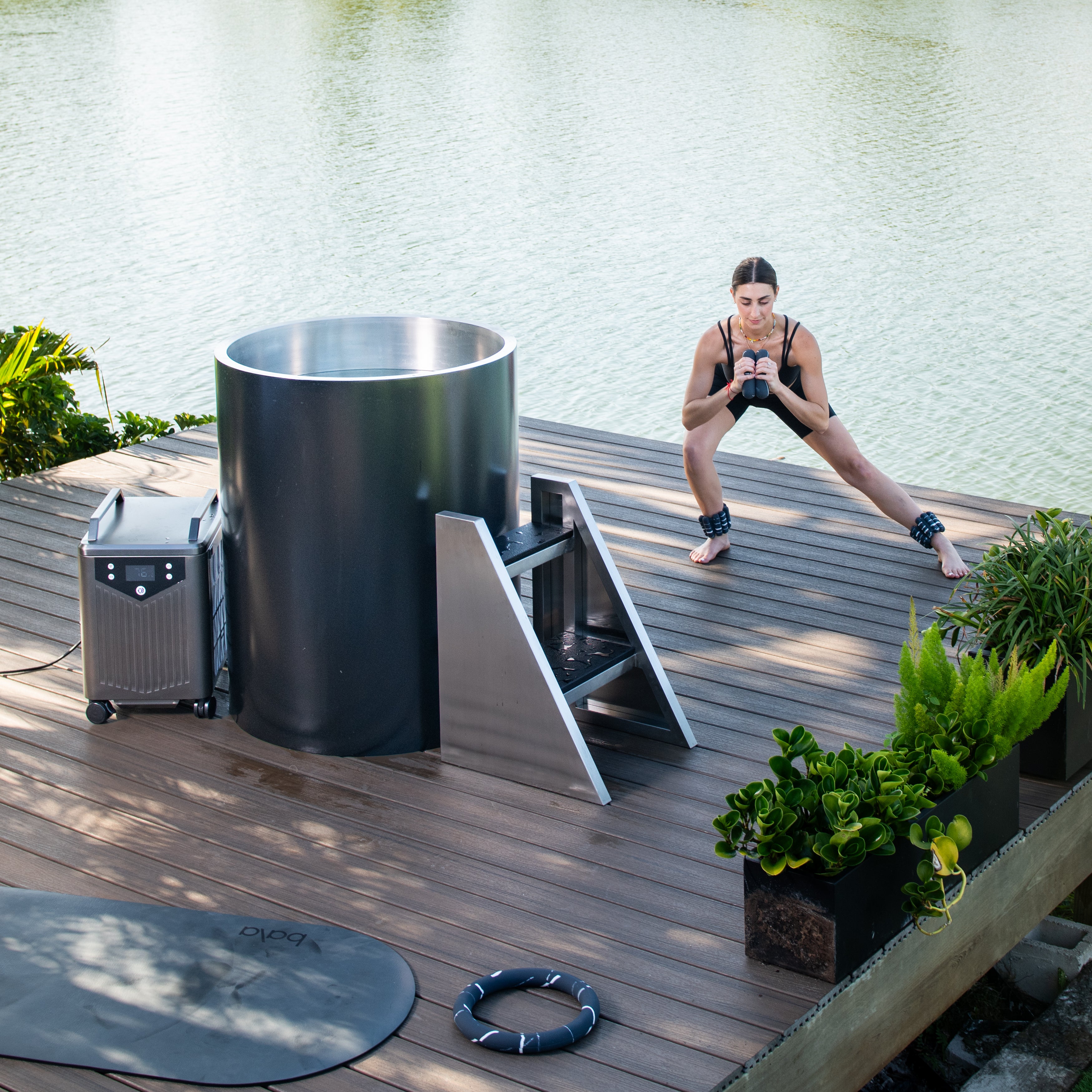 A woman in workout attire does side lunges with dumbbells on a wooden deck beside a Cyber Barrel cold plunge tub by a lake, surrounded by green plants and fitness equipment.