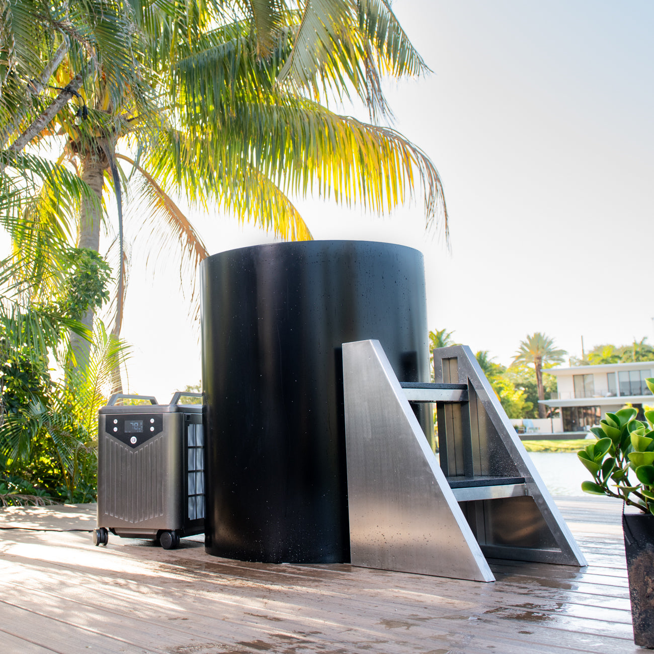 The Cyber Barrel, a black cylindrical cold plunge with vertical design and marine-grade stainless steel steps, sits on a wooden deck surrounded by palm trees, overlooking the waterfront and modern houses.