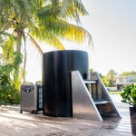 The Cyber Barrel, a black cylindrical cold plunge with vertical design and marine-grade stainless steel steps, sits on a wooden deck surrounded by palm trees, overlooking the waterfront and modern houses.