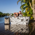 A man steps out of the Brainpod 2.0 acrylic cold plunge tub on a wooden deck beside a river, with palm trees, houses in the background, and a metal cooler nearby.