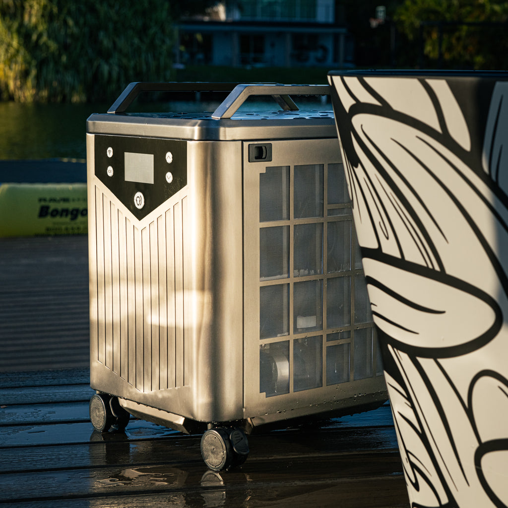 The Brainpod 2.0 Cold Plunge, a metal wheeled device with a digital display and vented sides, sits on a wooden deck near a large planter with artist-designed bold black-and-white leaf patterns as sunlight creates shadows and reflections.