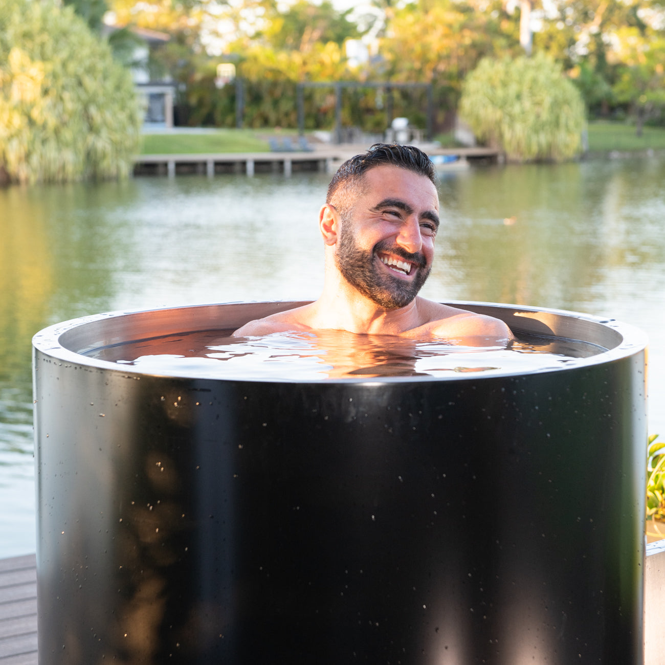 A smiling man relaxes in a Cyber Barrel Commercial Cold Plunge, enjoying a refreshing soak on a deck by a lake, surrounded by trees and lush greenery.