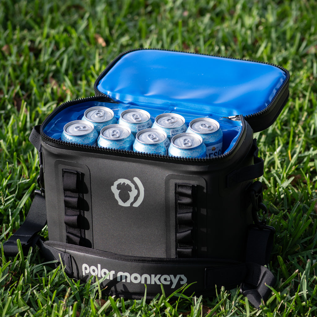 A black The Cave Cooler by Polar Monkeys sits open on grass, showing several drink cans inside its bright blue insulated interior, specially designed for excellent cold retention.