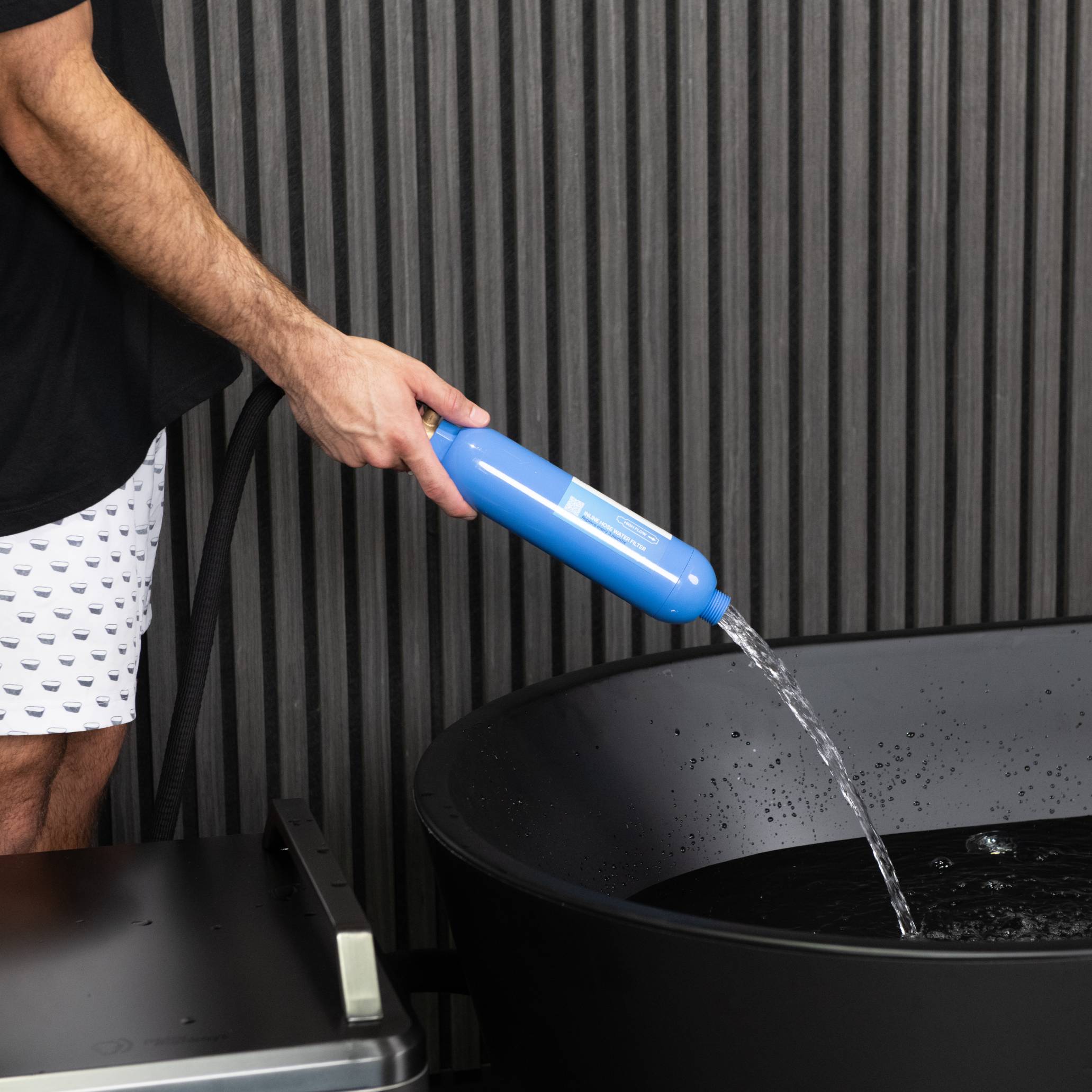 Wearing patterned shorts, a person uses the Hose Water Filter - 2 Pack to fill a black tub with water, set against vertical dark panels.