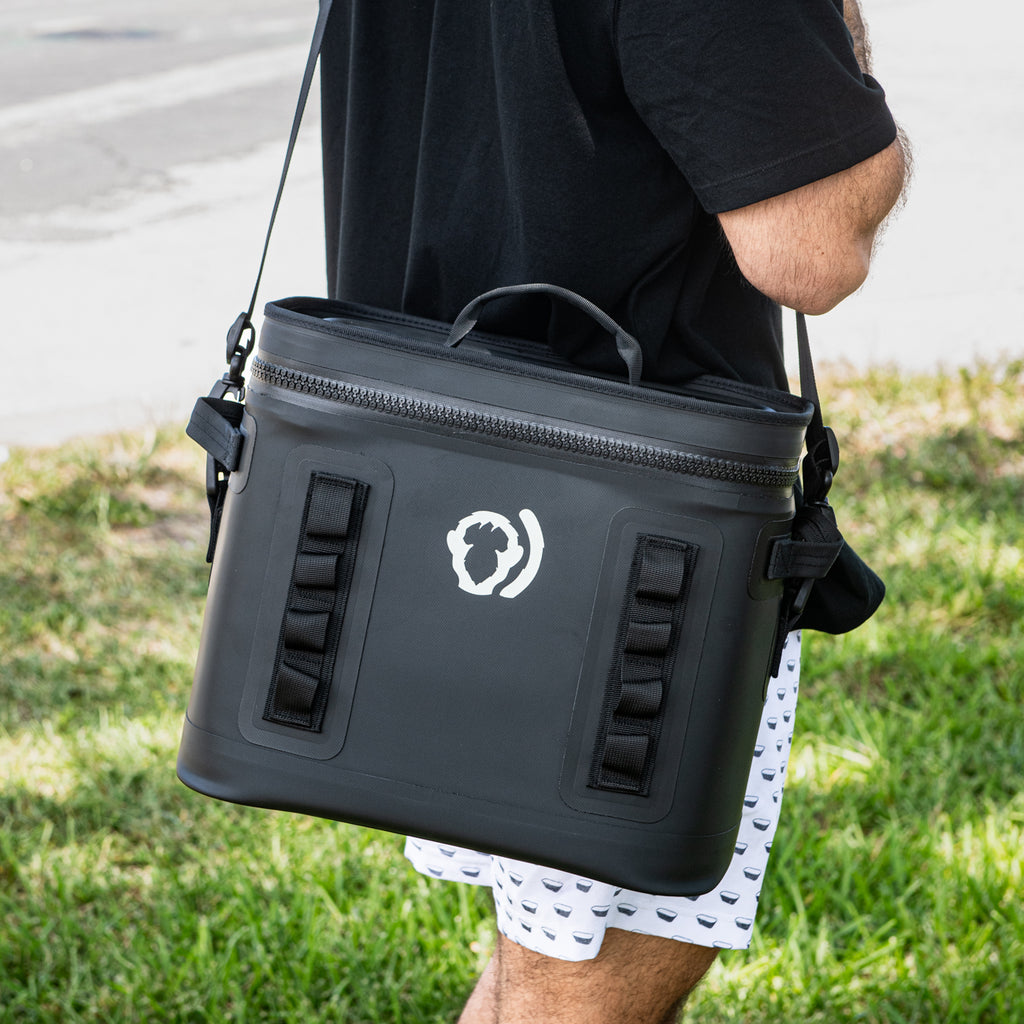 A person in a black shirt and patterned white shorts carries The Cave Cooler, a large, leak-proof black soft cooler bag with a handle and logo, while standing on green grass near a sidewalk.